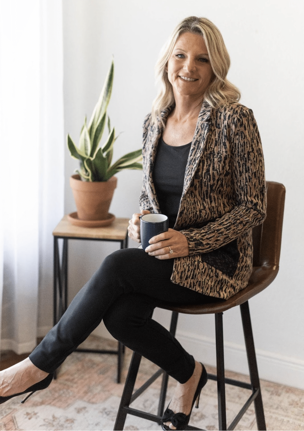 Ann Pearson sits and poses with a mug in her hand and a houseplant on a stool beside her.