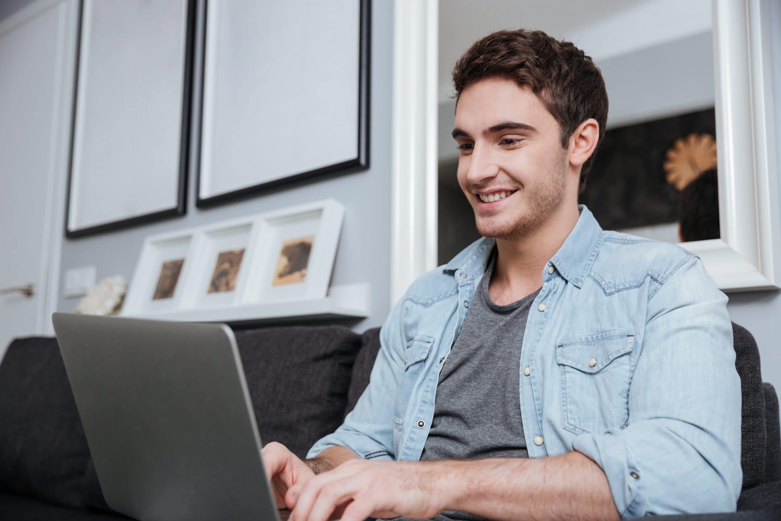 A young man smiling and working on his laptop while sitting on a couch in his living room