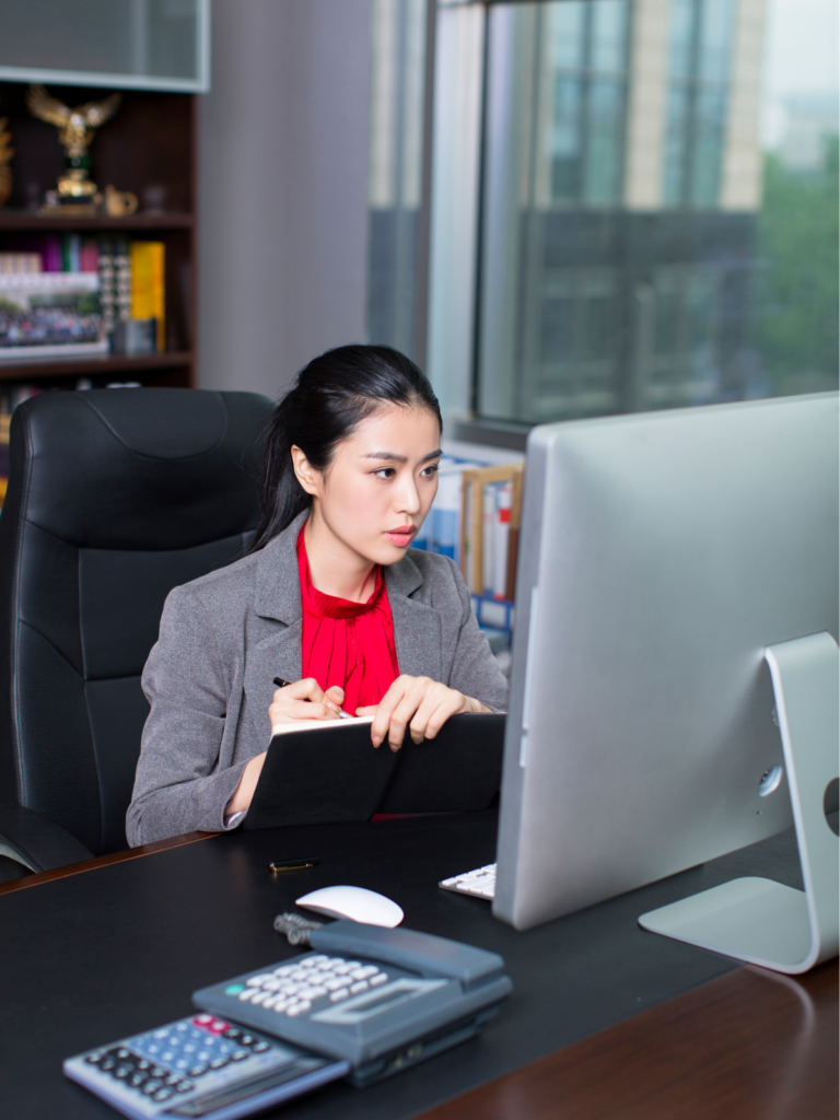 A paralegal manager in a grey suit and red top taking down details in her note from her desktop
