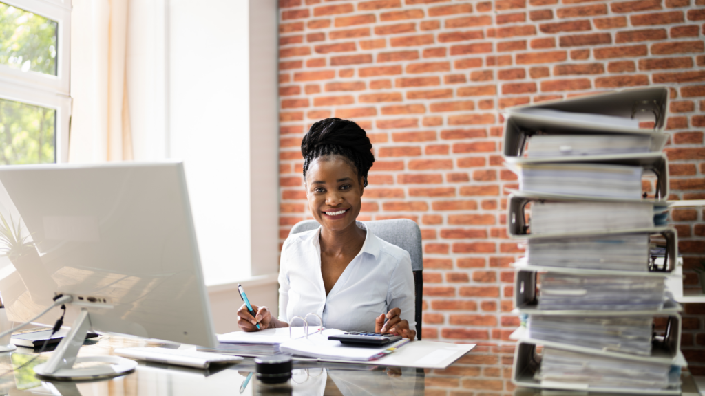 A paralegal sits at her desk with some huge stacked record files from which she has retrieved some documents.