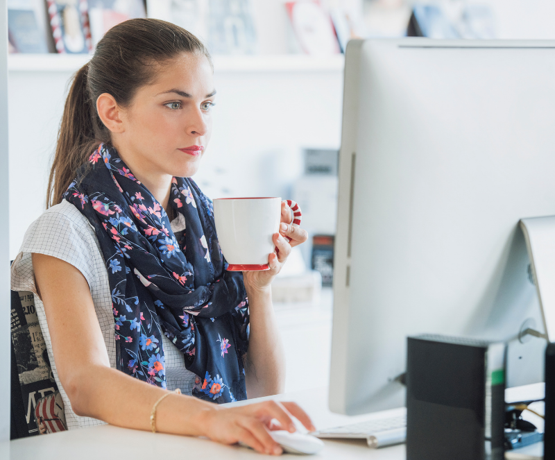 A woman with a beautiful flowery scarf around her neck working on her computer while holding her cup of coffee