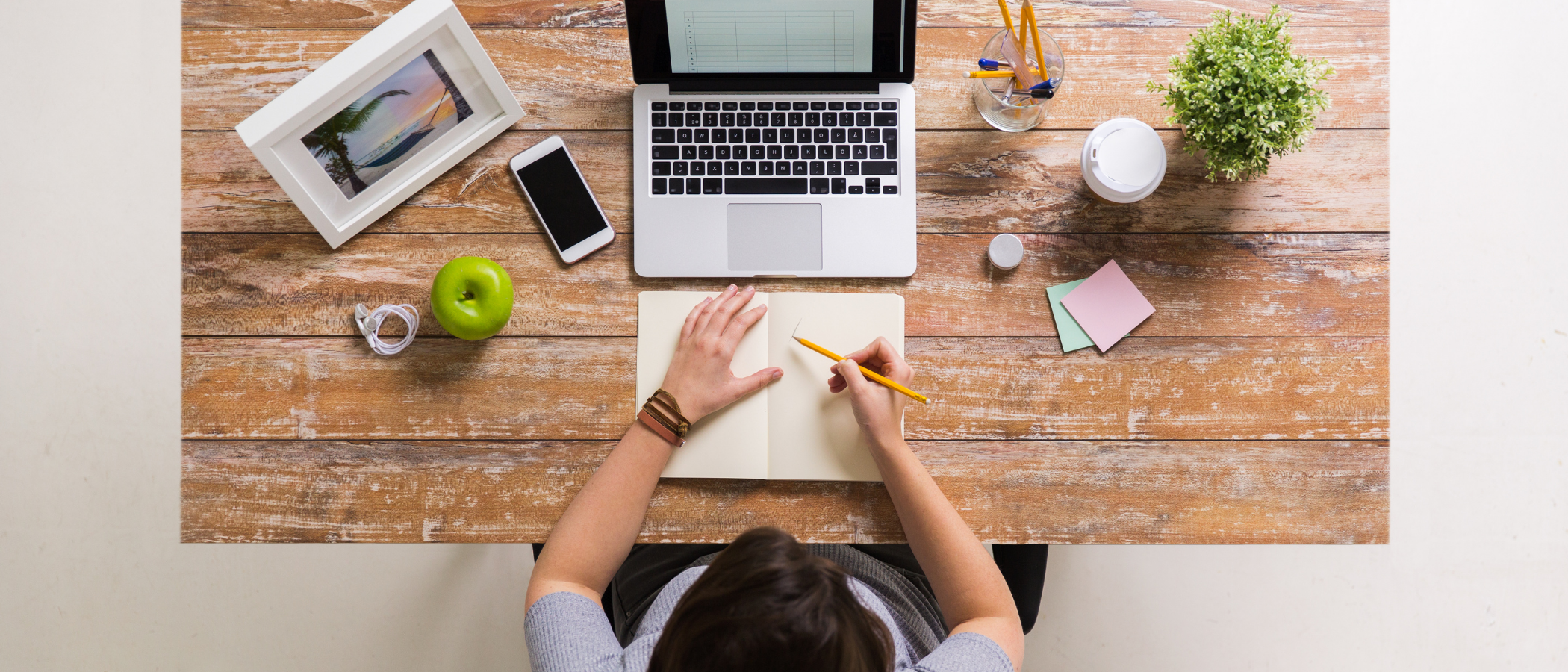 A woman working in a well-organized workspace with her note, picture frame, laptop, apple, coffee, and other stationery.