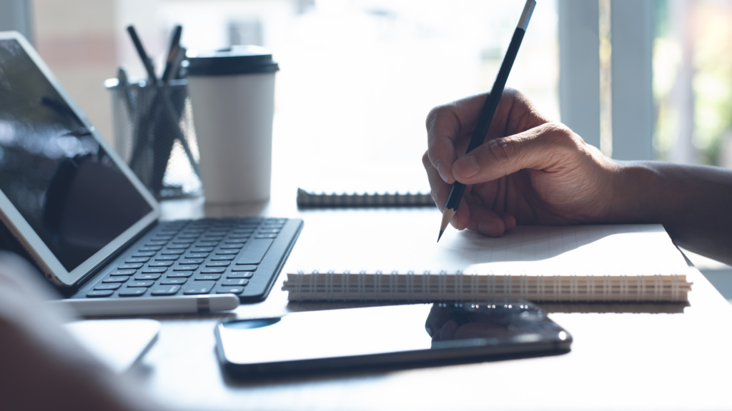 A trial paralegal's hand writing on a notepad with a PC, coffee, and phone on their working table.